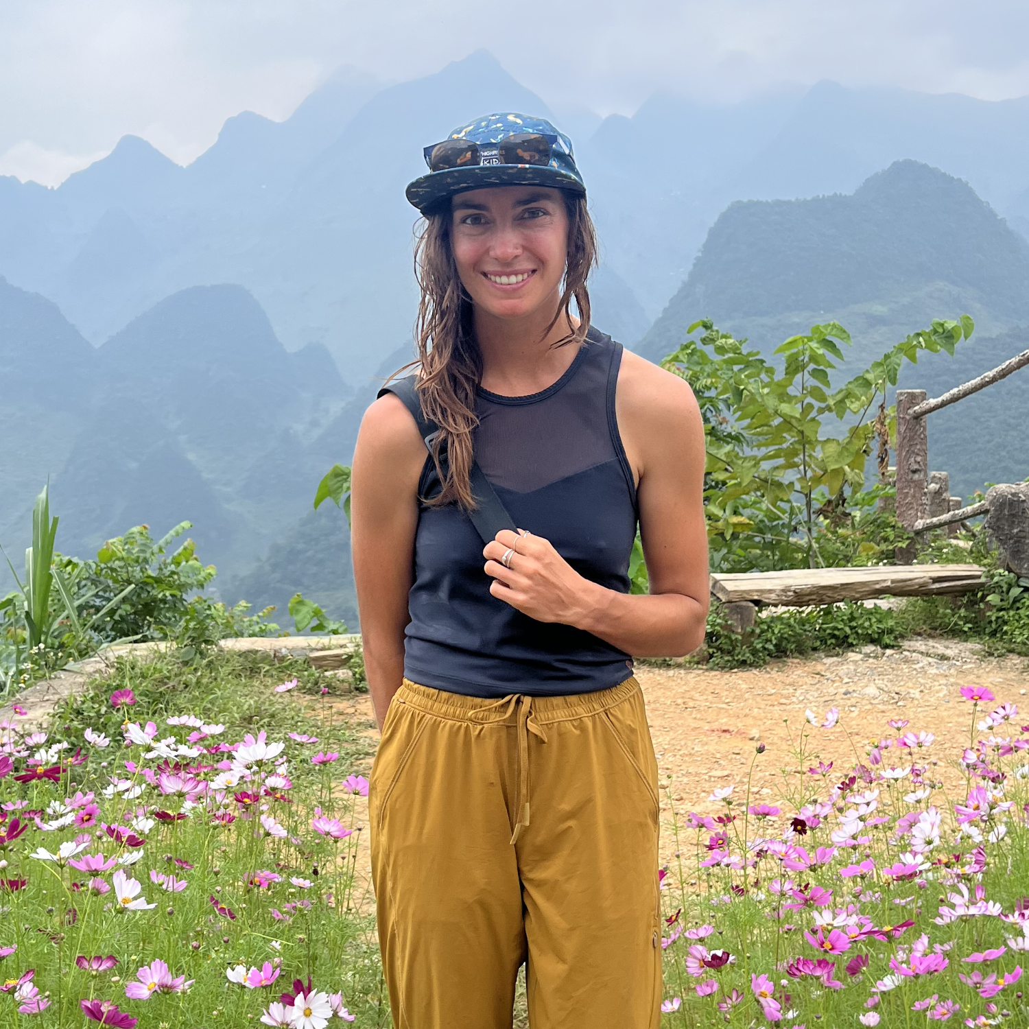 A woman in a blue hat, a black athletic shirt, and earthy yellow pants smiles for the camera while standing in a flower patch with a South American Mountain Range in the background.