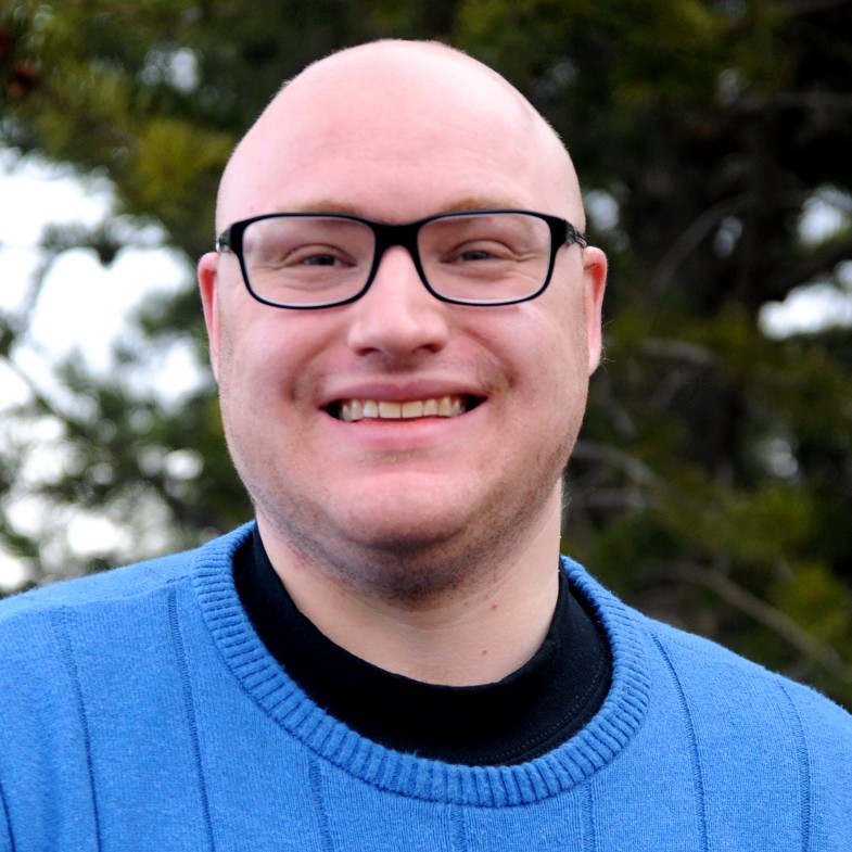 A smiling man standing outdoors in front of evergreen trees. He is wearing black glasses, a bright blue sweater, and a black shirt underneath. The background is softly blurred, giving a natural, forested feel.