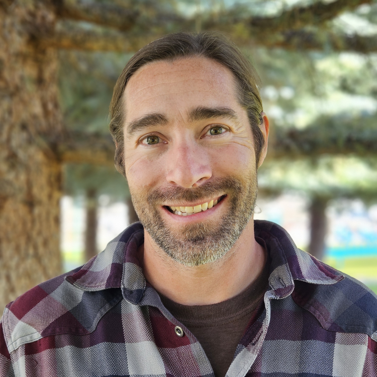Man smiling outdoors in front of pine trees, wearing a maroon and gray plaid shirt over a dark undershirt.