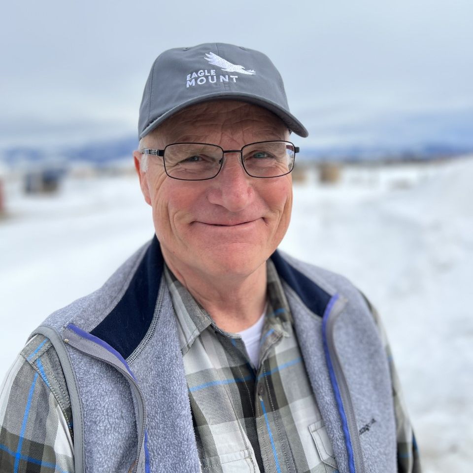 Older man smiling outdoors in snowy surroundings, wearing glasses, a gray Eagle Mount baseball cap, a plaid shirt, and a gray fleece vest.