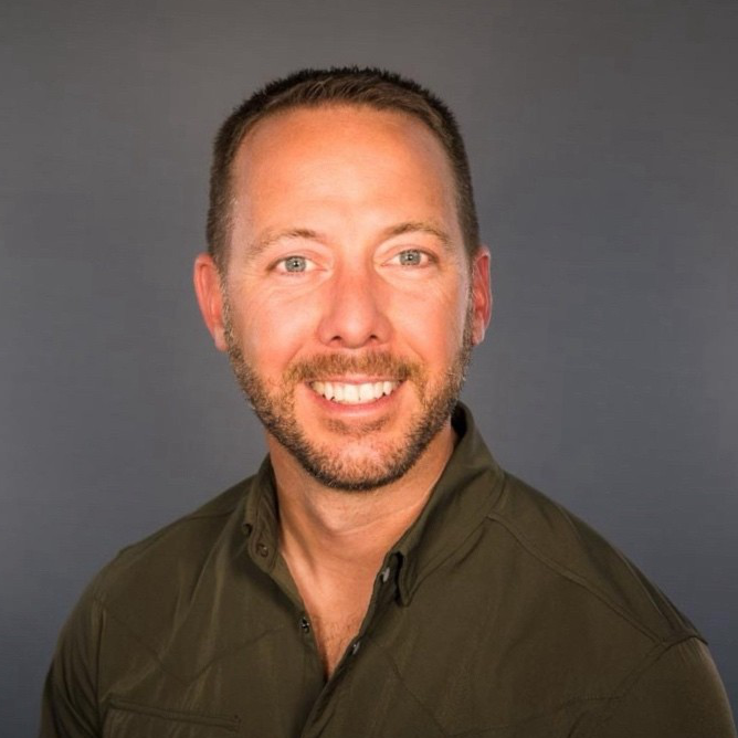 Smiling man with short brown hair and trimmed beard, wearing a dark green collared shirt, posed against a gray background.