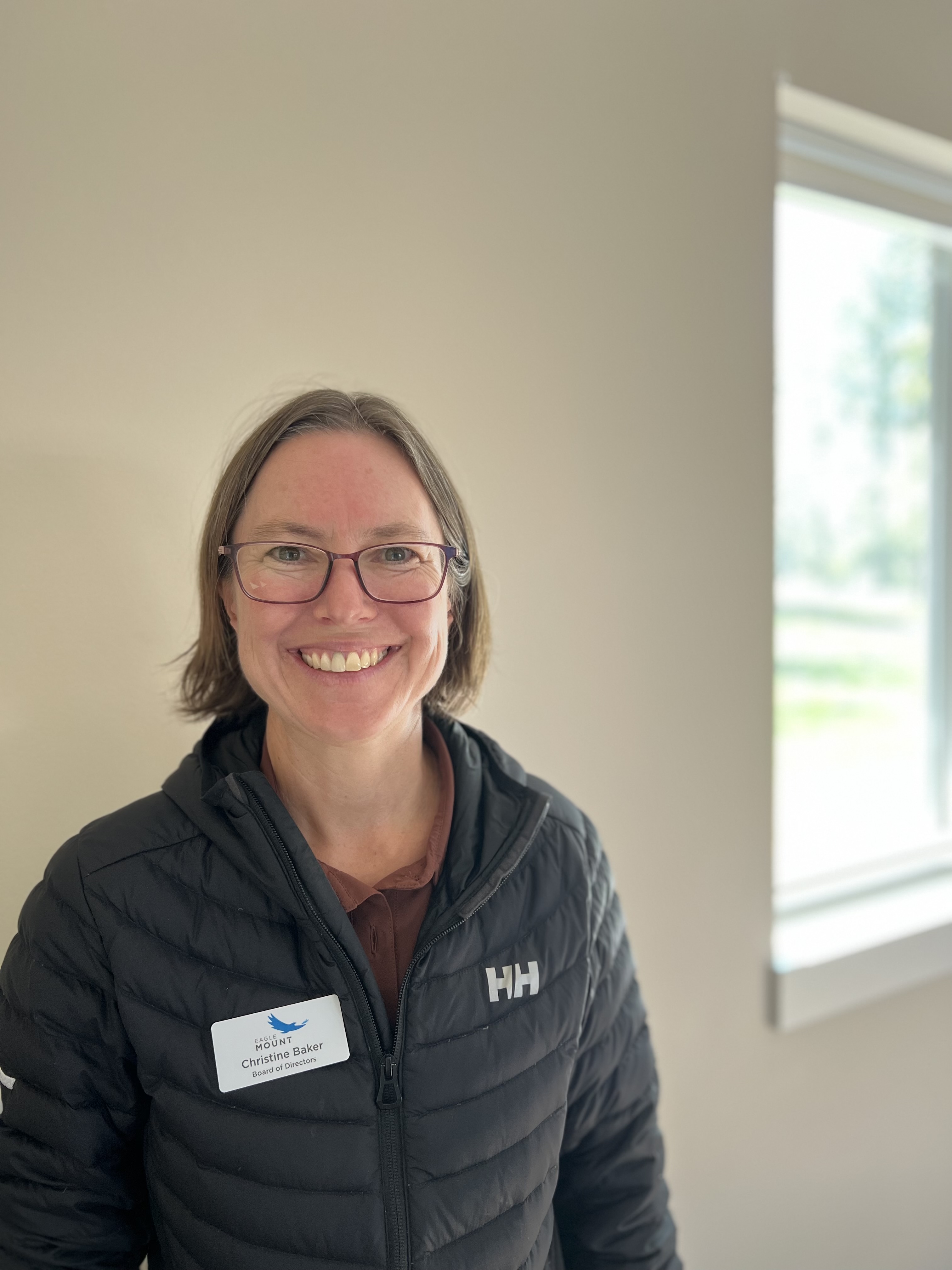Woman smiling indoors near a sunlit window, wearing glasses, a black Helly Hansen jacket, and a name tag that reads 'Christine Baker – Board of Directors.'