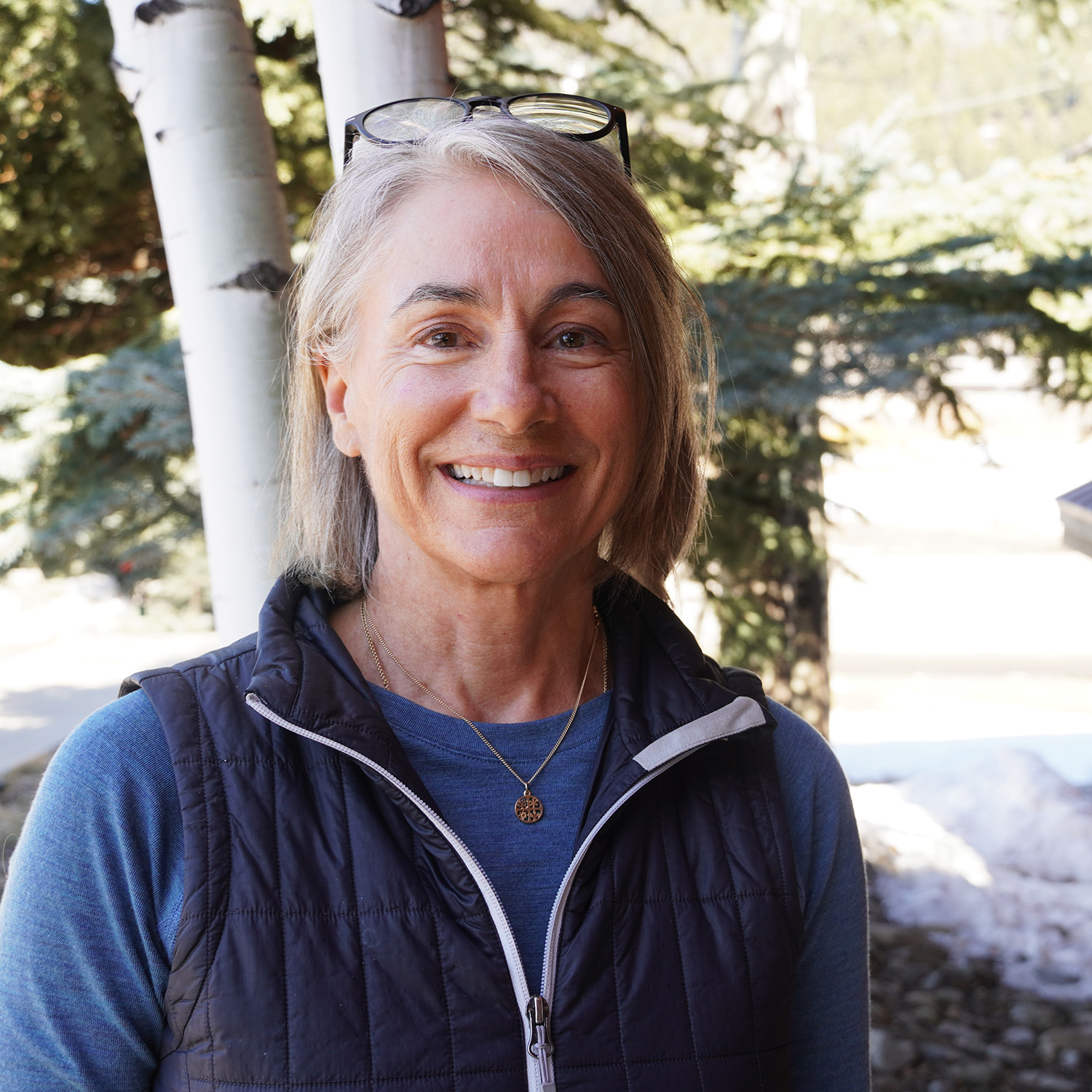 Woman smiling outdoors in front of evergreen trees and birch trunks, wearing a blue shirt and black vest with glasses resting on her head.
