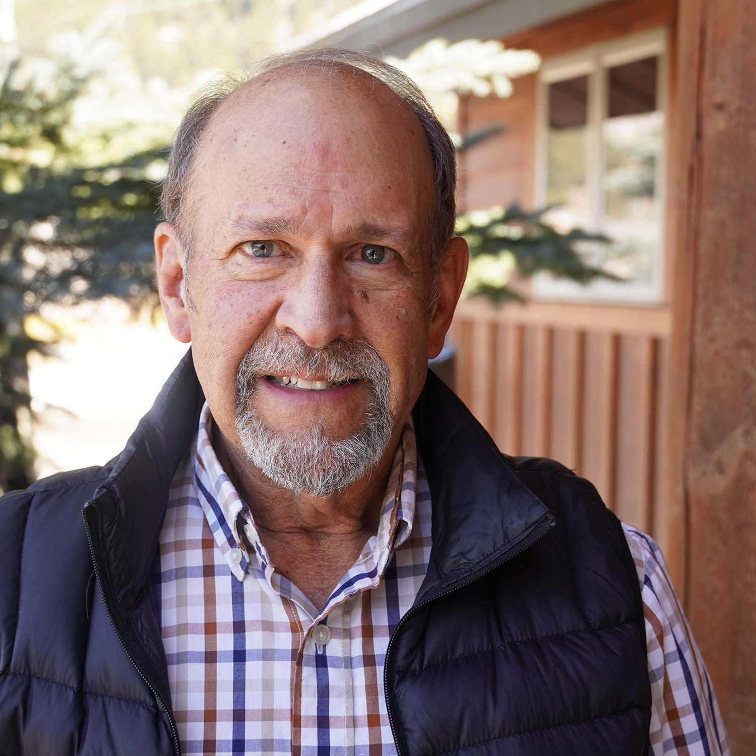 Smiling older man with short gray hair and a trimmed beard, wearing a plaid button-up shirt and a black puffer vest, standing outside near a wooden building with trees in the background.