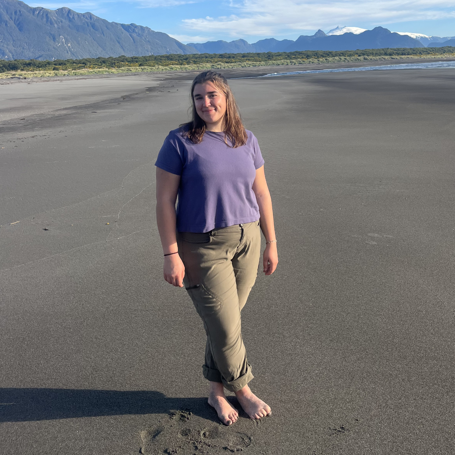 Woman standing barefoot on a wide, dark-sand beach with her legs crossed, wearing a purple shirt and khaki pants, with forested foothills and snow-capped mountains in the background under a blue sky.