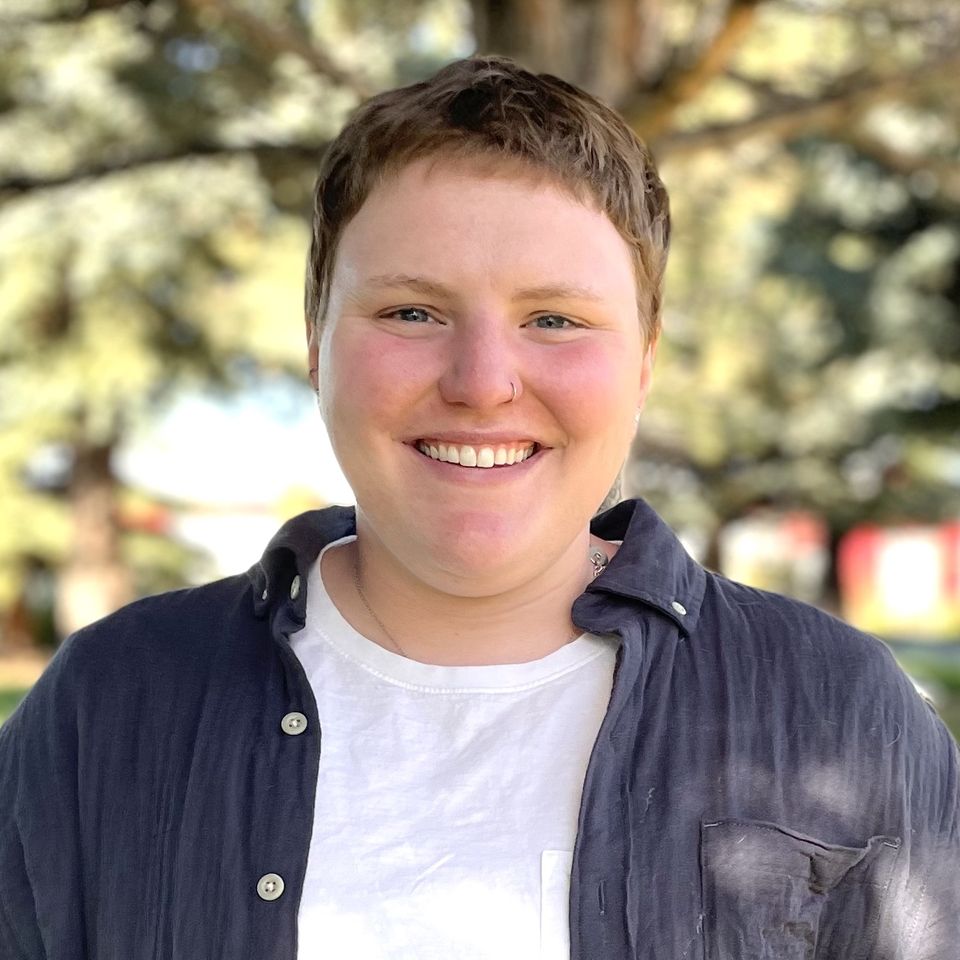 Finn Connors smiles outdoors with sunlight filtering through the trees behind them. They are wearing a white T-shirt and a dark button-up shirt, with short hair and a nose ring.