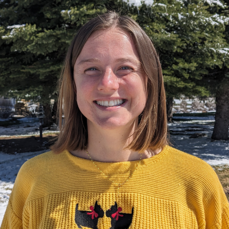 Woman smiling outdoors on a sunny winter day, wearing a yellow sweater with two black dog silhouettes and red bows on the front. Snow and evergreen trees in the background.