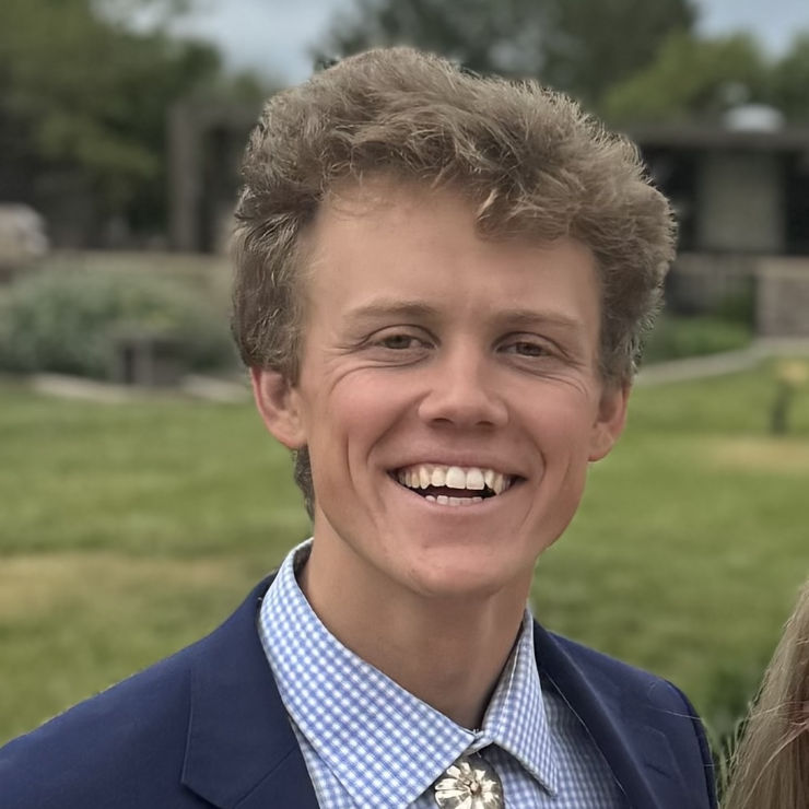 A young man wearing a blue blazer and checkered shirt smiles outdoors with greenery and buildings in the background.