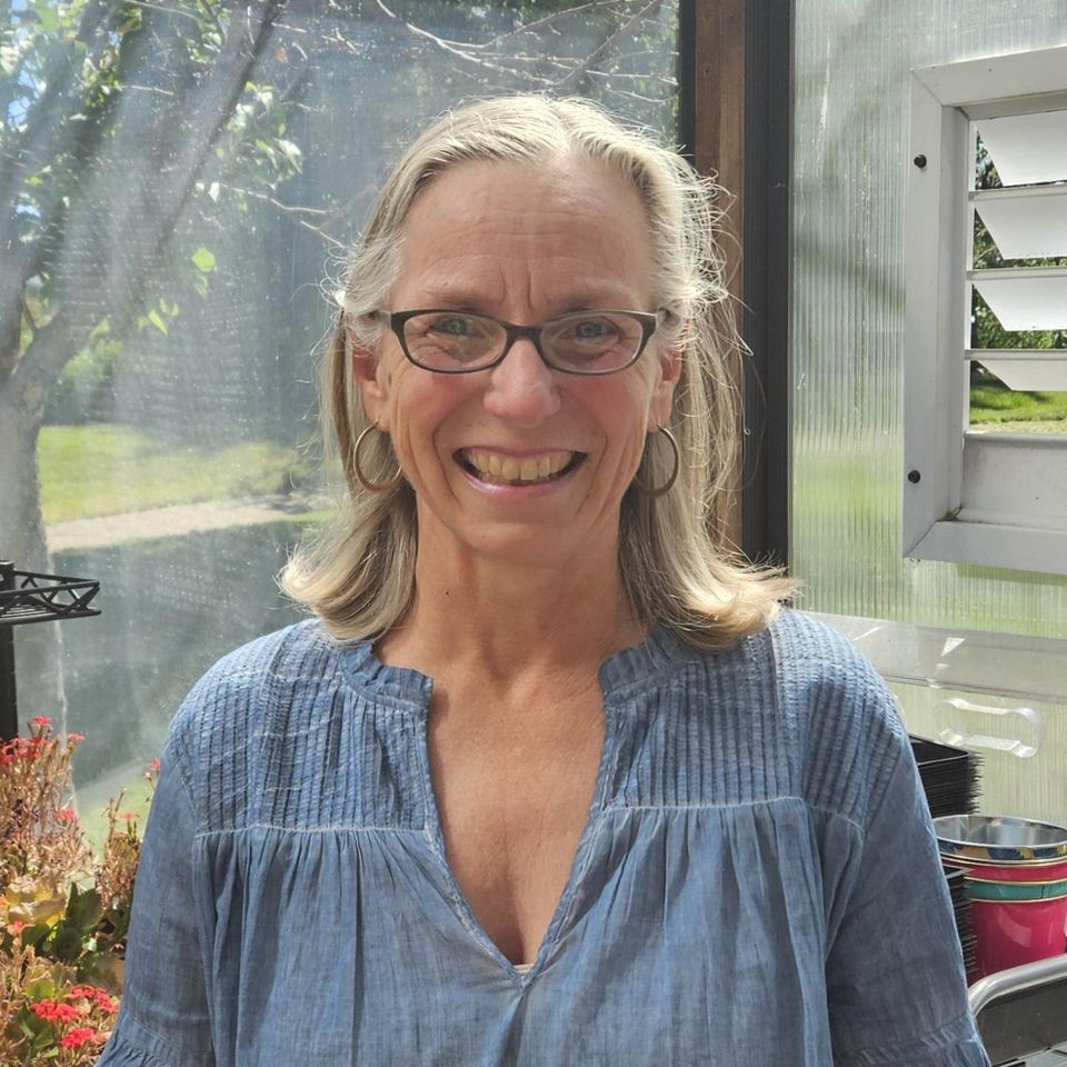 Smiling woman with shoulder-length gray-blonde hair and glasses, wearing hoop earrings and a light blue pleated blouse, standing in a sunlit greenhouse with potted plants in the background.