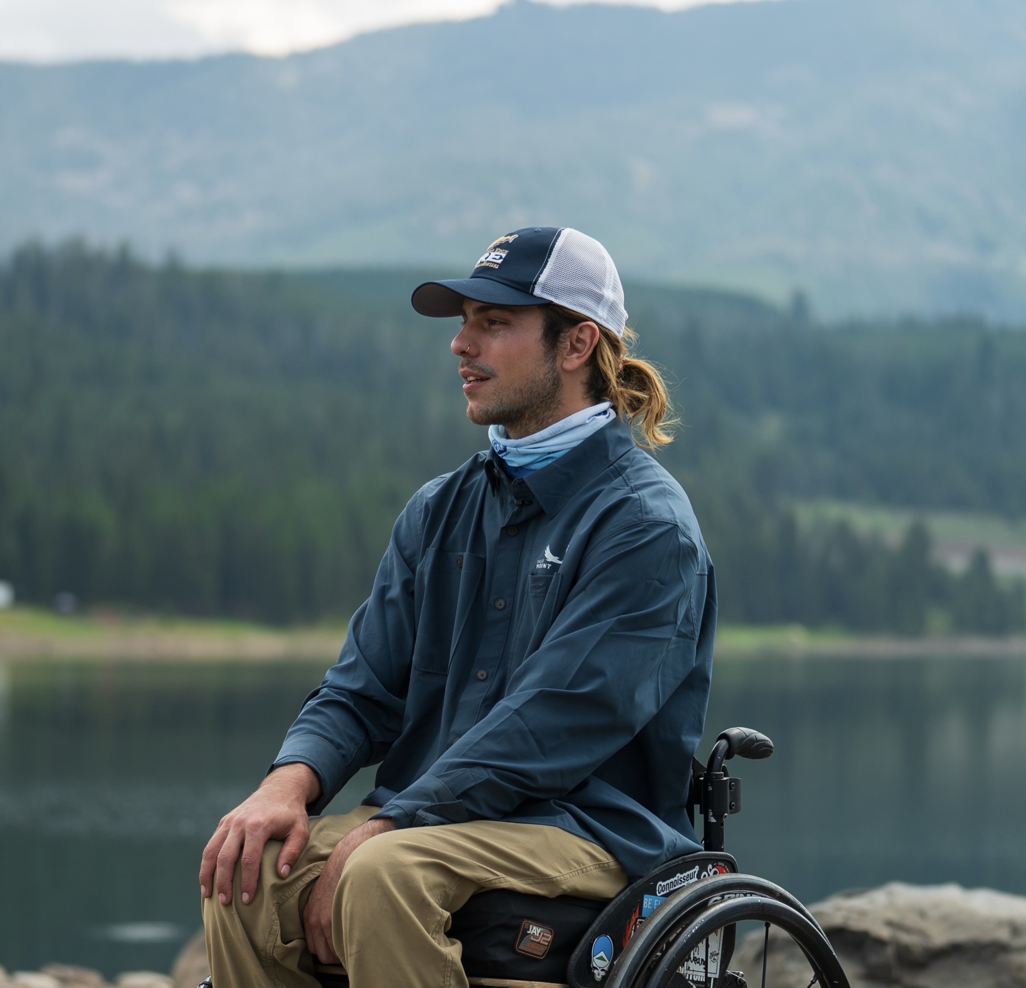 A man sitting in a wheelchair outdoors near a calm lake, with forested hills and mountains in the background. He has a ponytail and is wearing a blue fishing shirt, tan pants, a trucker hat, and a neck gaiter. His wheelchair has various stickers on it. The sky is overcast and the scene is peaceful.