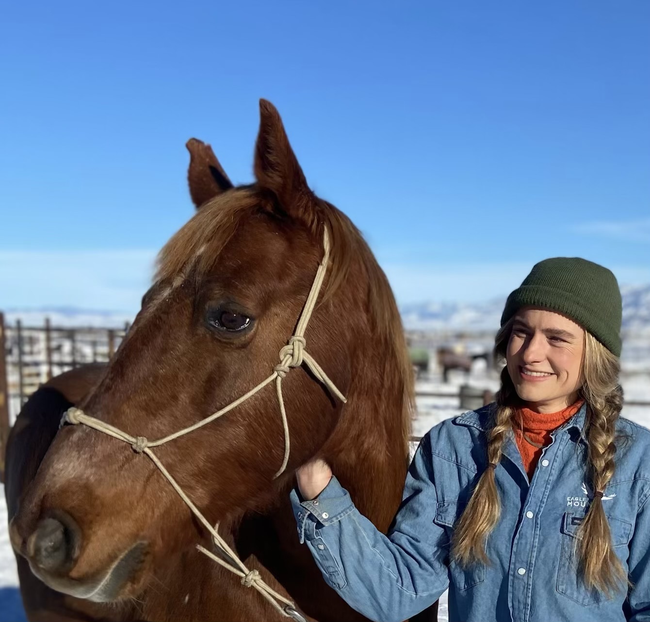 A woman wearing a green beanie, denim Eagle Mount jacket, and orange scarf smiles while standing next to a brown horse with a rope halter. Snow covers the ground and mountains are visible in the background.