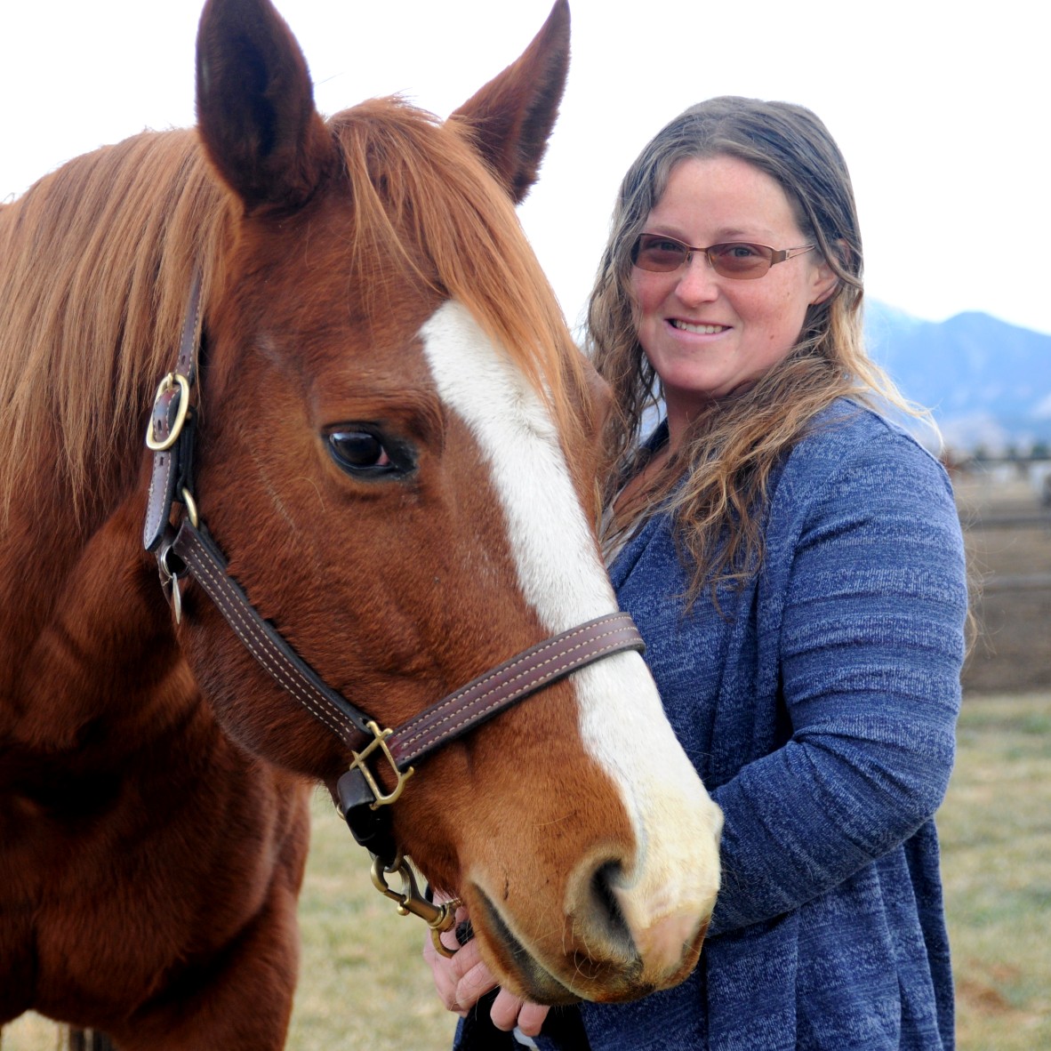 Smiling woman wearing glasses and a blue sweater stands beside a chestnut horse with a white blaze on its face. They are outdoors with a mountain in the background.