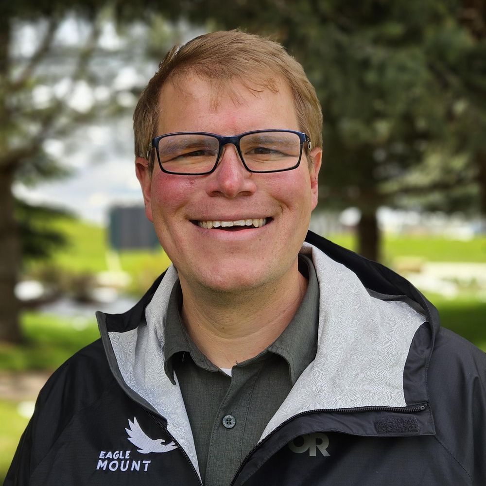 Man smiling outdoors, wearing glasses and a black Eagle Mount jacket with a light gray lining, standing in front of blurred green trees and a grassy background.