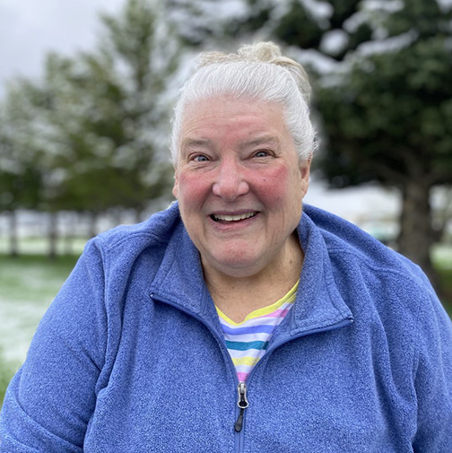 Portrait of a smiling older woman with white hair pulled back in a bun, wearing a bright blue fleece jacket over a striped shirt. She is outdoors in front of a green lawn and trees, with light snow visible on the grass and branches.