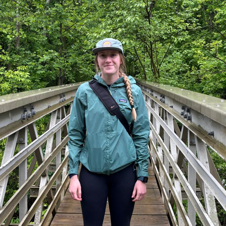 Young woman standing on a wooden footbridge in a lush forest, wearing a green rain jacket, black leggings, a crossbody bag, and a light green cap with a braided ponytail over one shoulder.