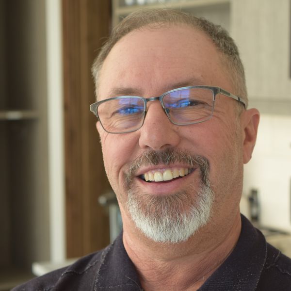 A smiling man with short hair, glasses, and a gray goatee wearing a dark collared shirt, indoors with blurred background.