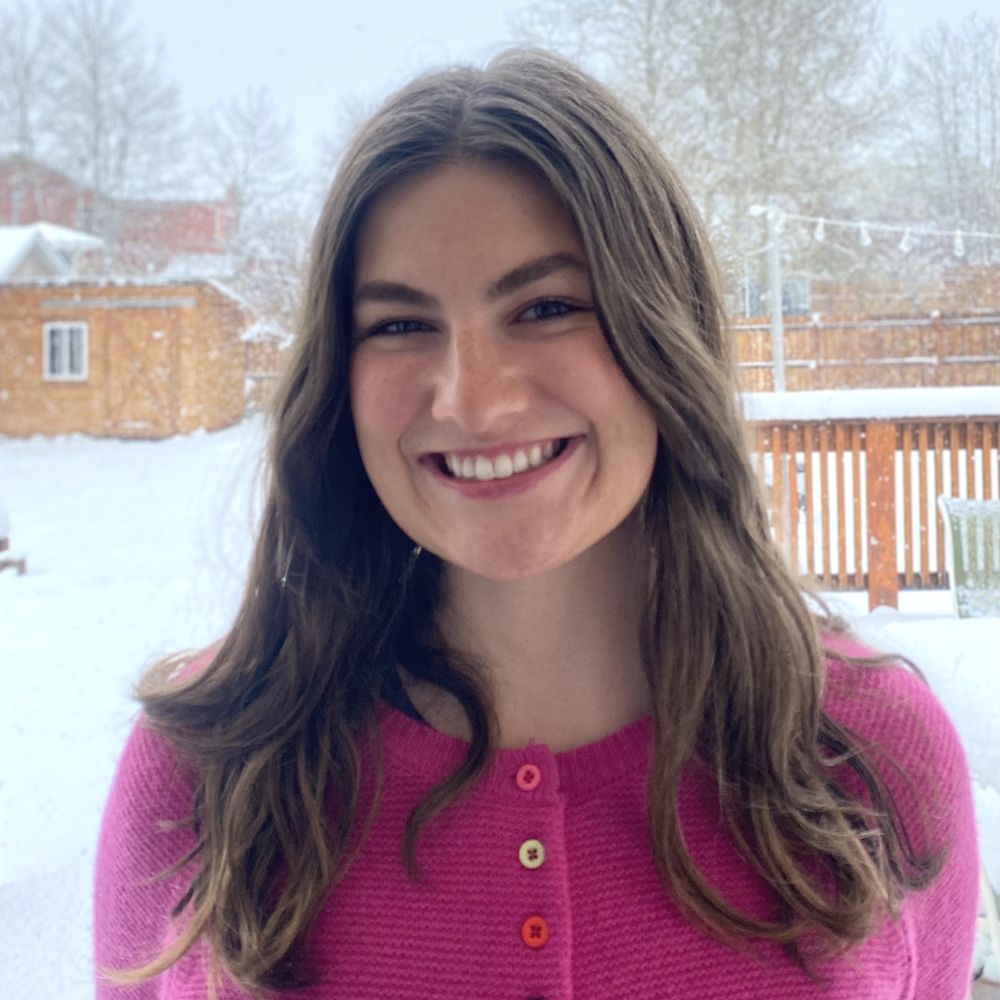 Woman smiling in front of a snowy backyard, wearing a bright pink sweater with colorful buttons.