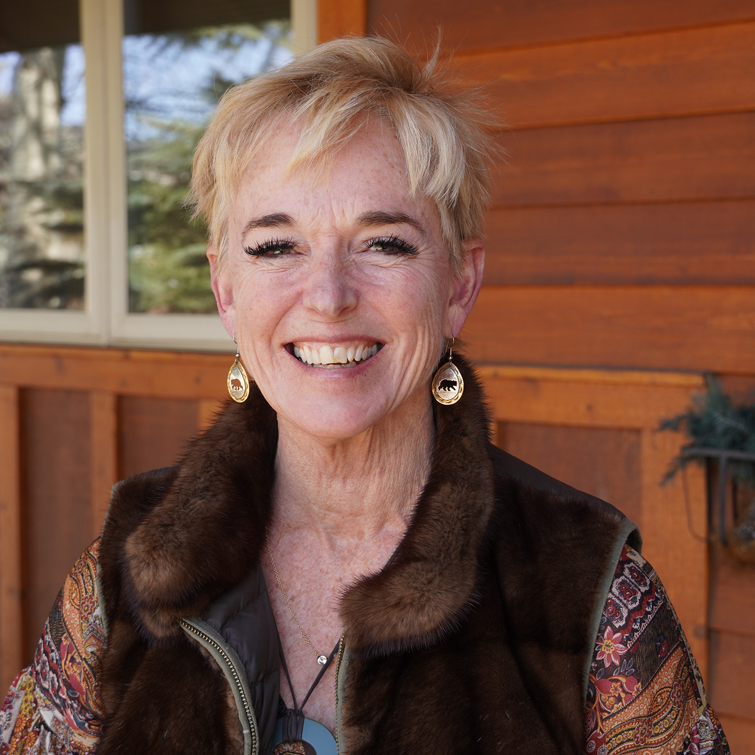 Smiling woman with short blonde hair, wearing patterned earrings, a brown fur-trimmed vest, and a colorful patterned top, standing outside in front of a wooden building.
