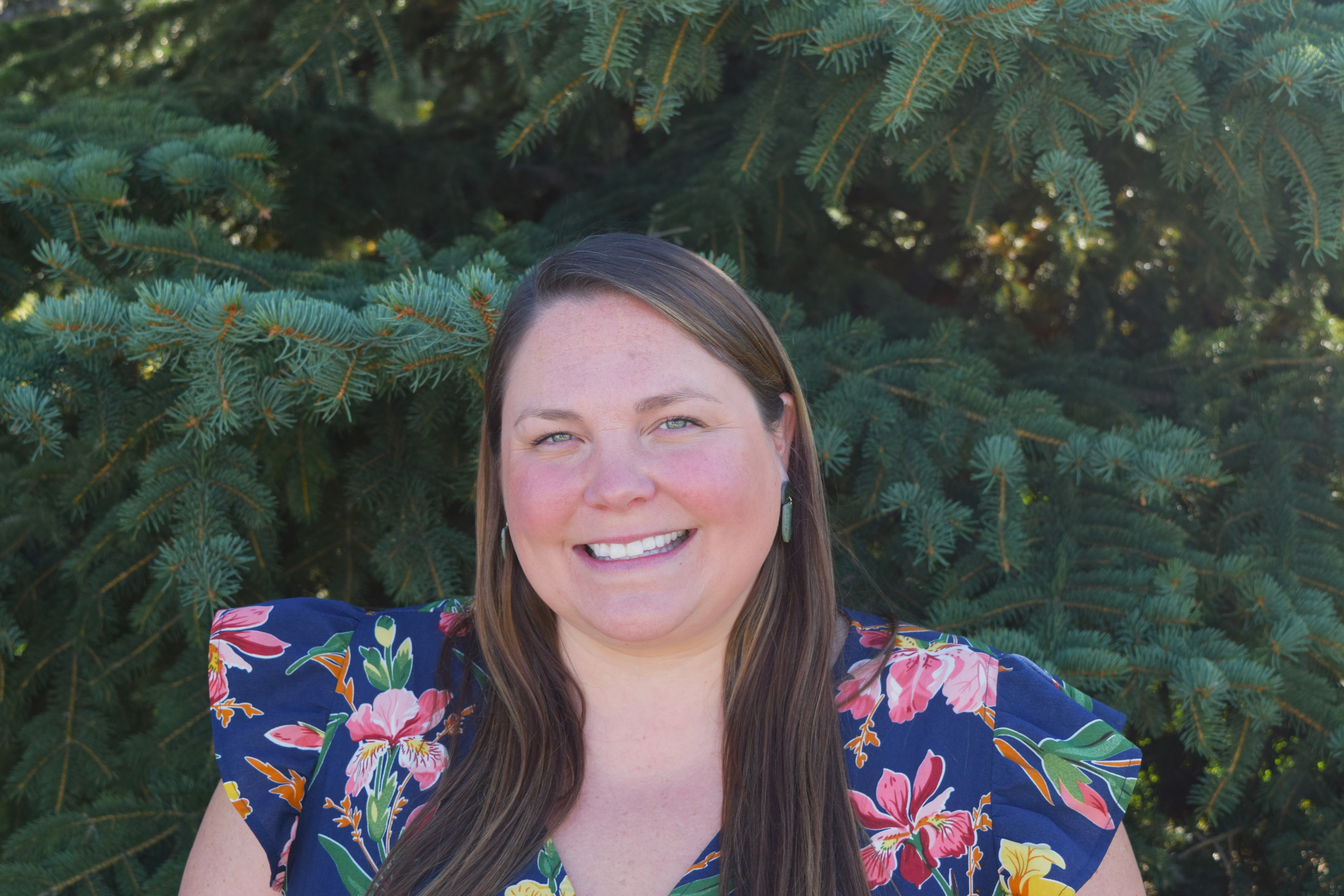 Woman smiling in front of evergreen trees, wearing a navy blue floral top with pink, orange, and green flowers.