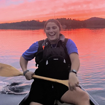Smiling woman paddling a canoe at sunset on a calm lake, wearing a blue shirt and black life vest, with silhouetted hills and a vibrant pink and orange sky in the background.