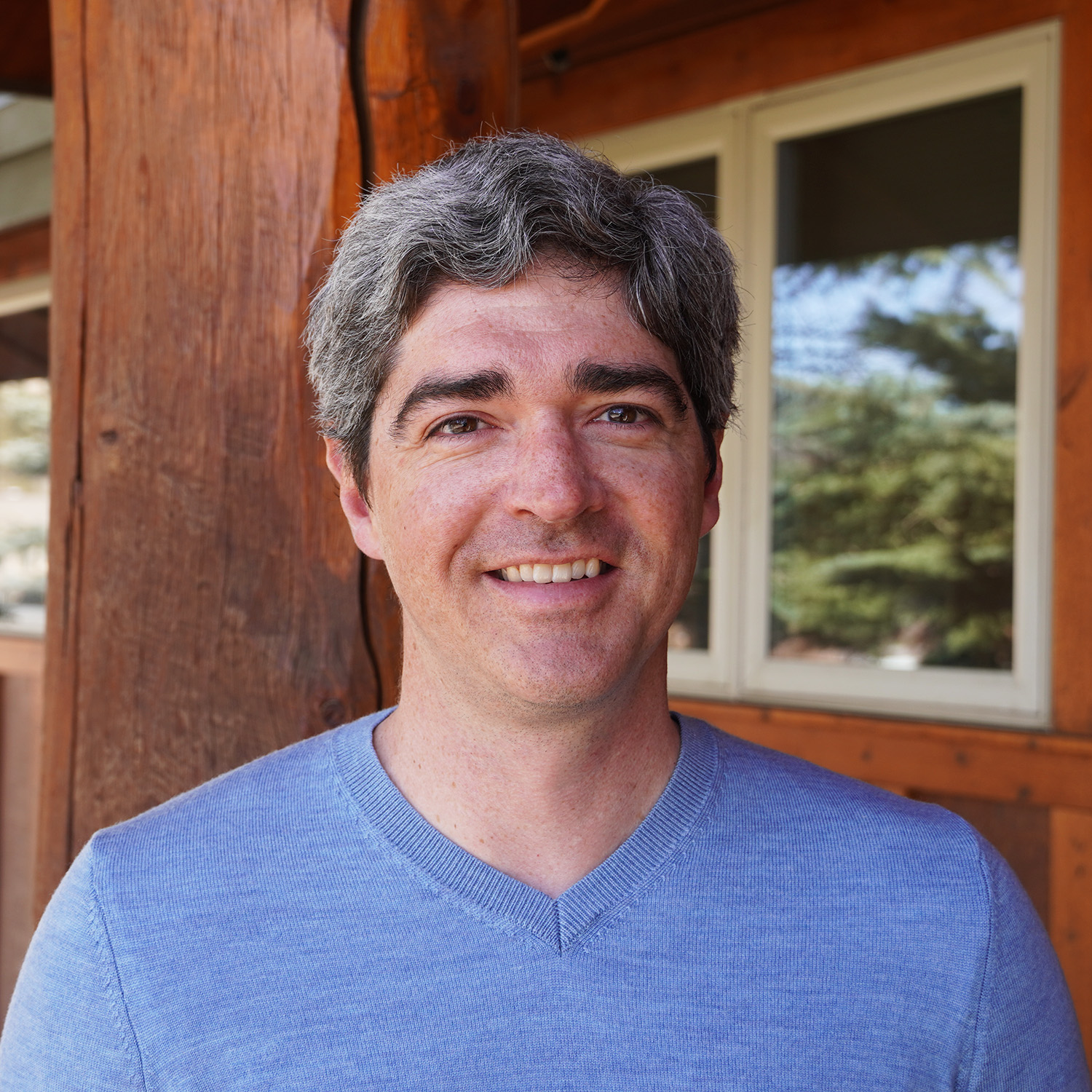 Portrait of a smiling man with short gray hair, wearing a light blue V-neck sweater. He is standing outside near a wooden building with windows and trees reflected in the glass.