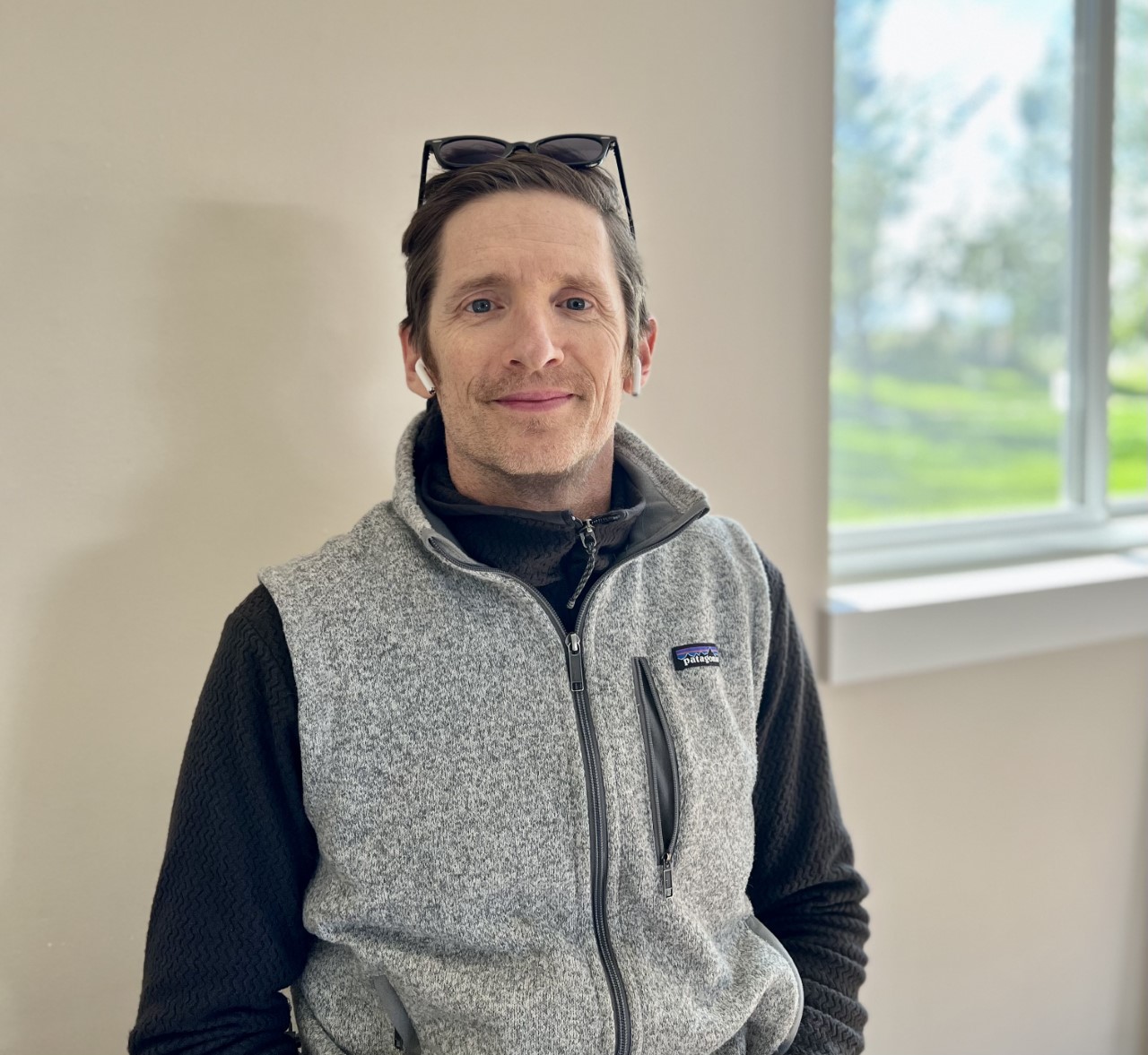 Portrait of a man seated indoors, wearing a gray fleece vest over a black zip-up sweater. He has short brown hair, wireless earbuds, and sunglasses resting on his head. A bright window with trees and grass outside is in the background.