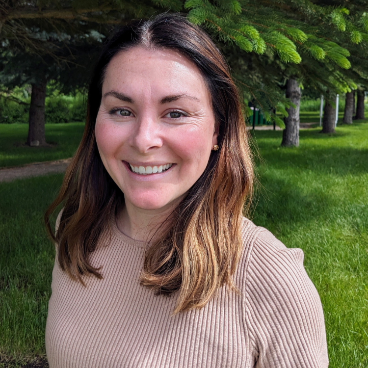 Smiling woman standing outside on a sunny day, wearing a ribbed beige sweater. She has shoulder-length brown hair with highlights and is surrounded by green grass and pine trees.