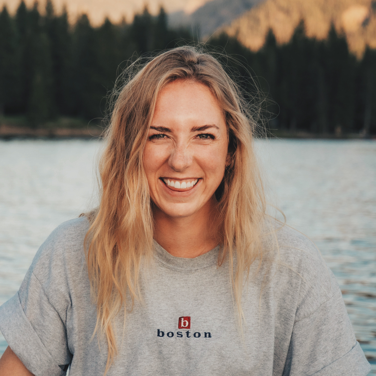 Smiling woman with long blonde hair wearing a gray T-shirt that says “Boston,” standing outdoors in front of a calm lake with pine trees and mountains in the background.