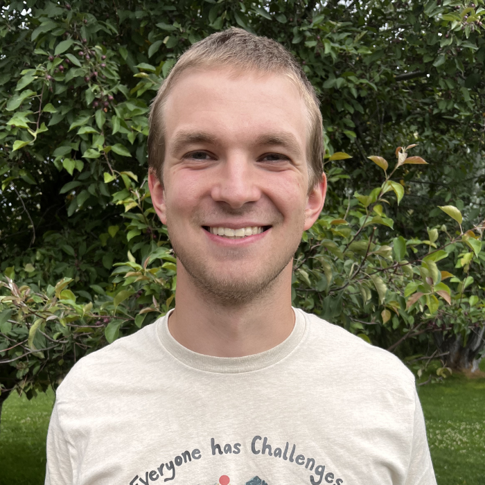 Man smiling outdoors in front of leafy trees, wearing a light-colored T-shirt that says 