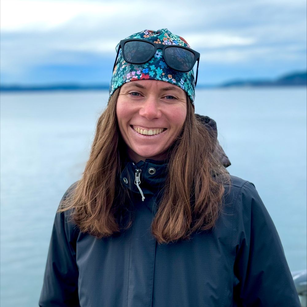 A smiling woman standing near a large body of water on a cloudy day. She has long brown hair and is wearing a dark jacket and a colorful floral-patterned headband with sunglasses resting on top. The calm water and distant shoreline create a serene coastal backdrop.