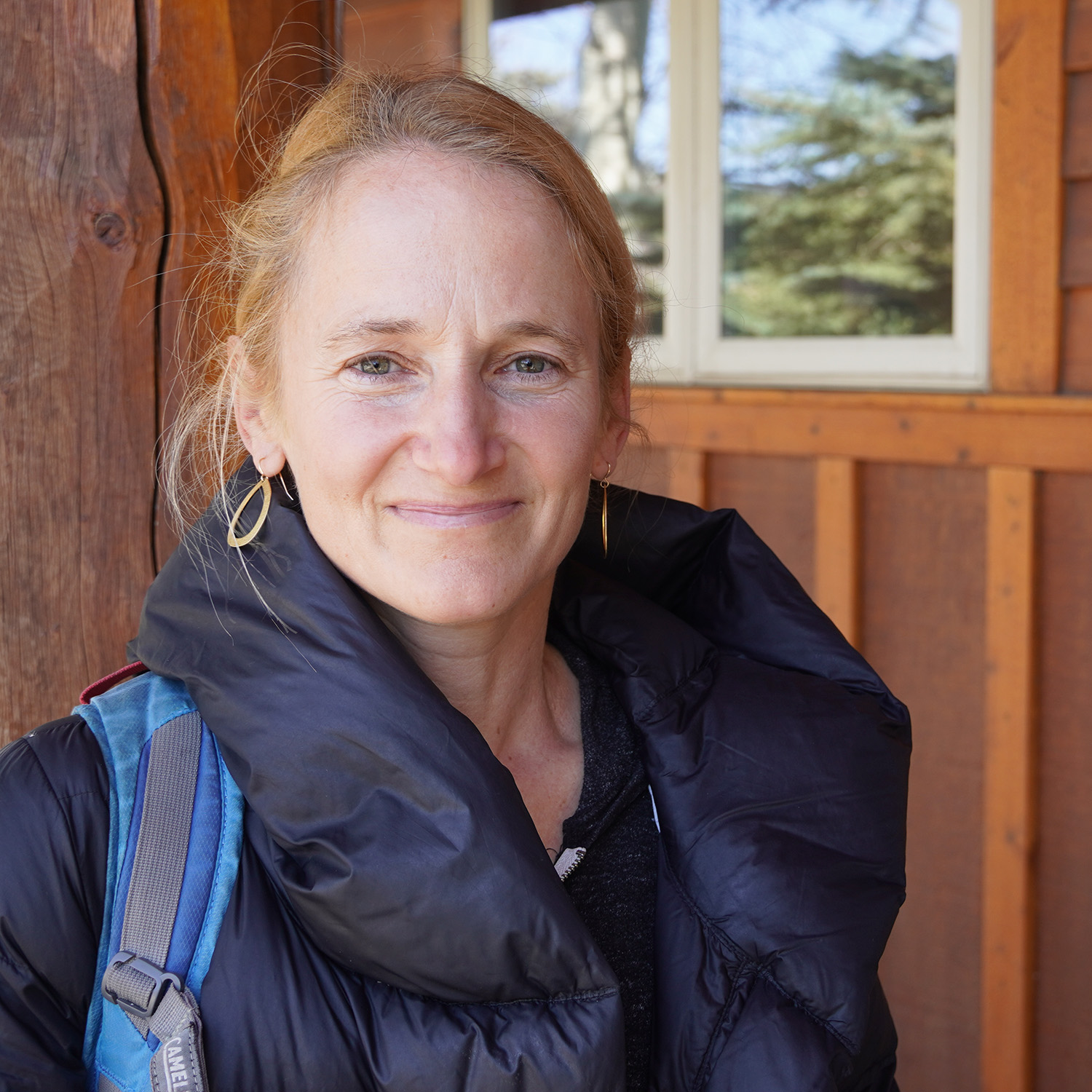 A woman with light hair pulled back, wearing gold hoop earrings and a black puffy jacket with a backpack strap visible, smiling slightly in front of a wooden building with a window.