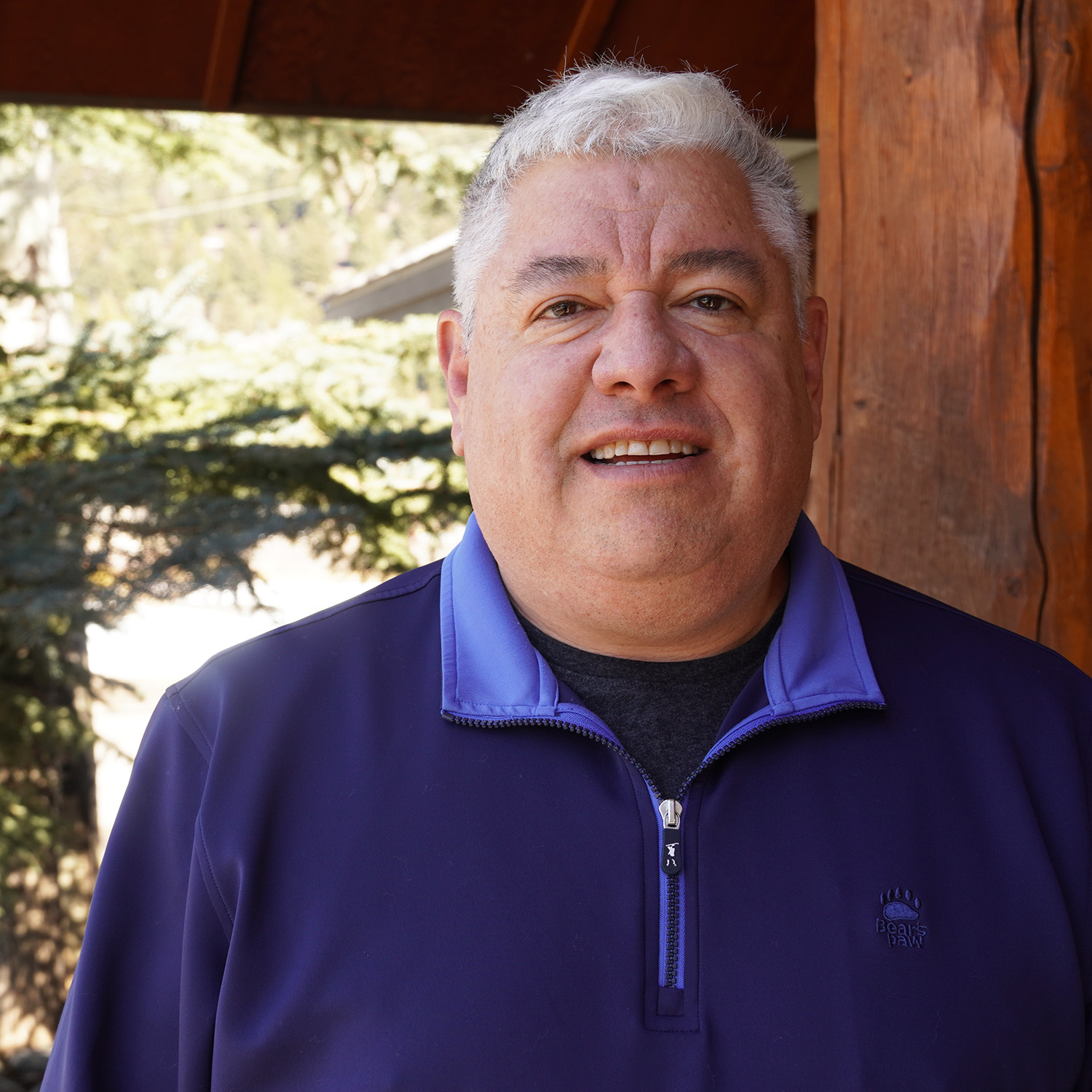 Man smiling outdoors on a sunny day, standing beside a wooden pillar and wearing a dark blue quarter-zip jacket.