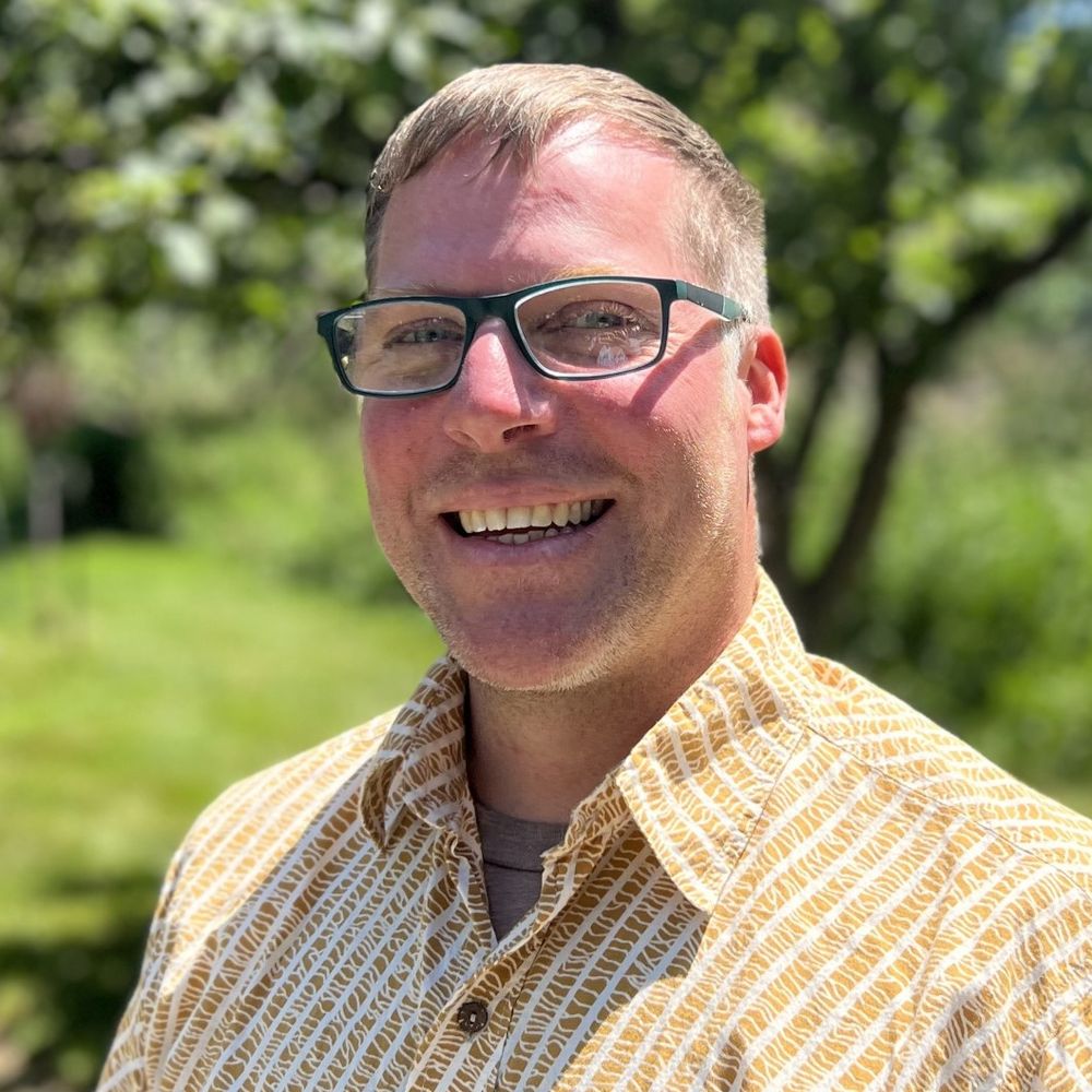 Man smiling outdoors on a sunny day, wearing dark-rimmed glasses and a patterned yellow shirt. Green trees and grass fill the background.