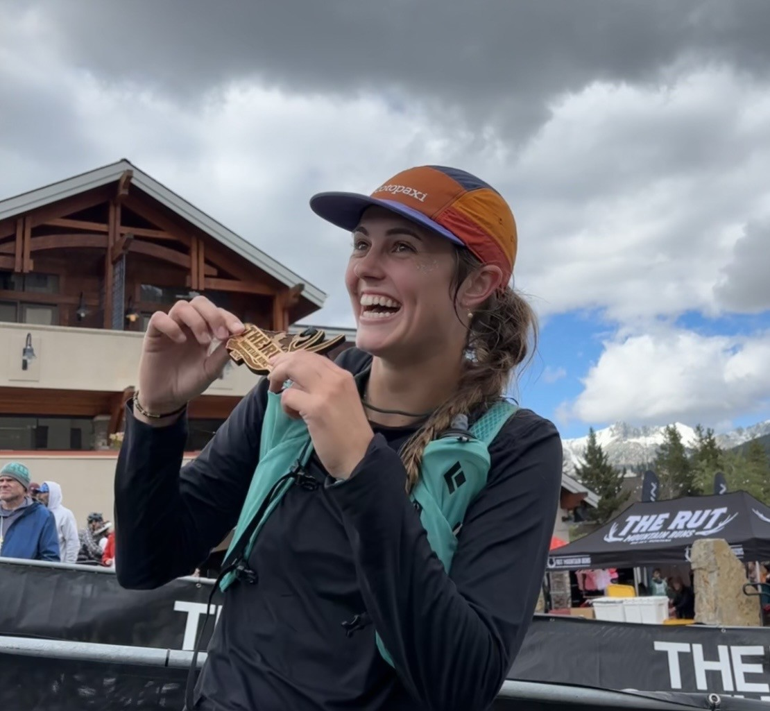 Smiling woman wearing a multicolored cap and hydration vest holds a race medal near her mouth at an outdoor event, with a lodge, banners reading “The Rut,” and snow-capped mountains in the background.