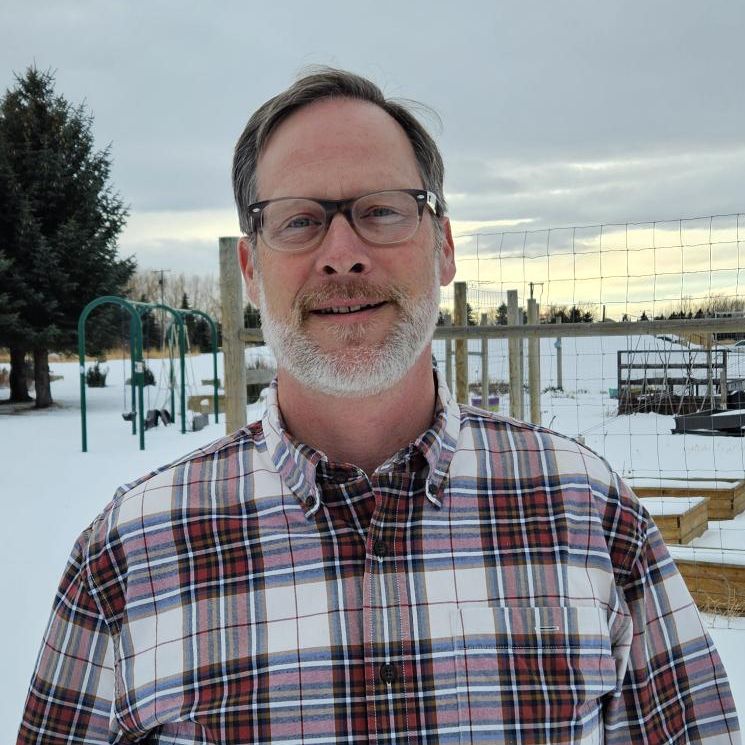 Man with glasses and a graying beard standing outside in a snowy landscape, wearing a red and white plaid shirt. Behind him are trees, a fence, and raised garden beds under a cloudy winter sky.
