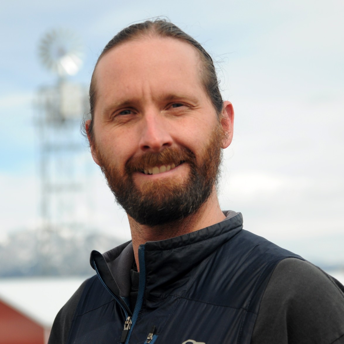 Smiling man with long brown hair tied back and a full beard, wearing a dark zip-up jacket, standing outdoors with a blurred windmill structure and cloudy sky in the background.