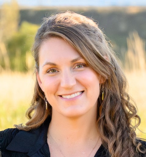 Smiling woman with long, wavy brown hair swept to one side, wearing a black top and dangling earrings, standing outdoors in a sunlit field with tall grasses and blurred hills in the background.